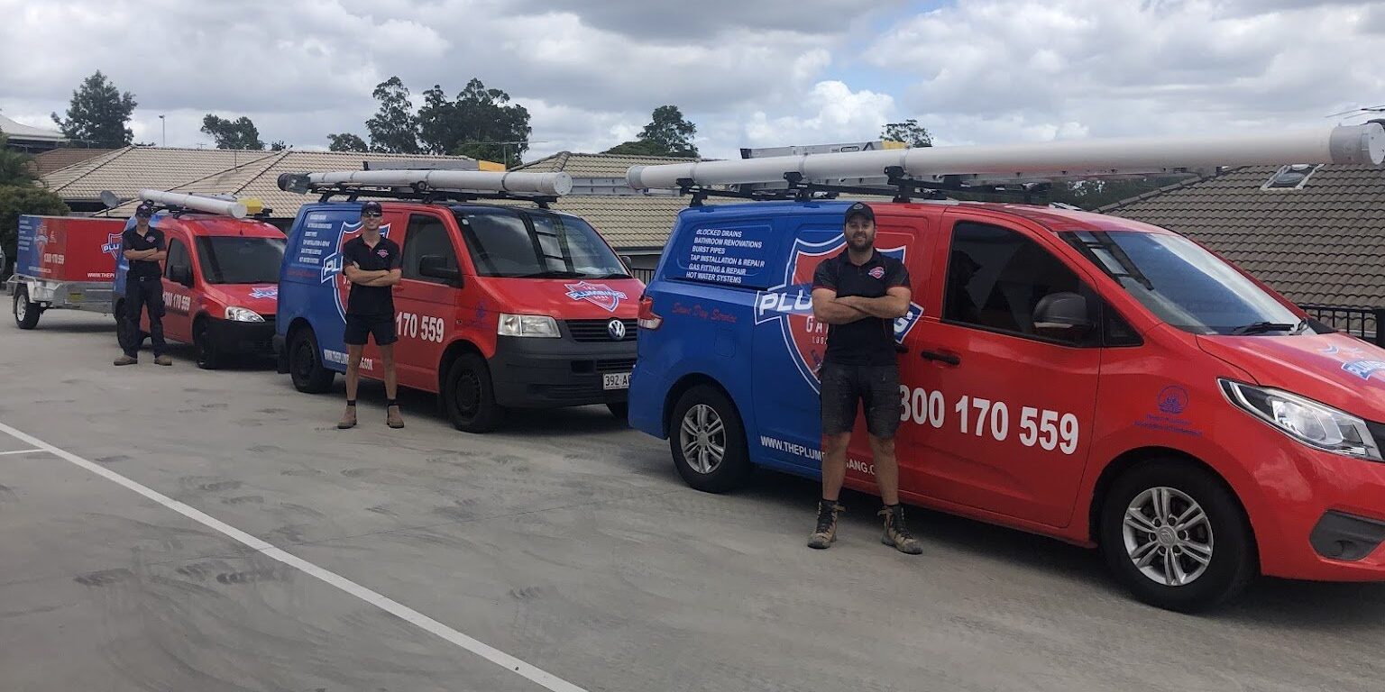 Three plumbers stood in line next to their The Plumbing Gang branded vehicles